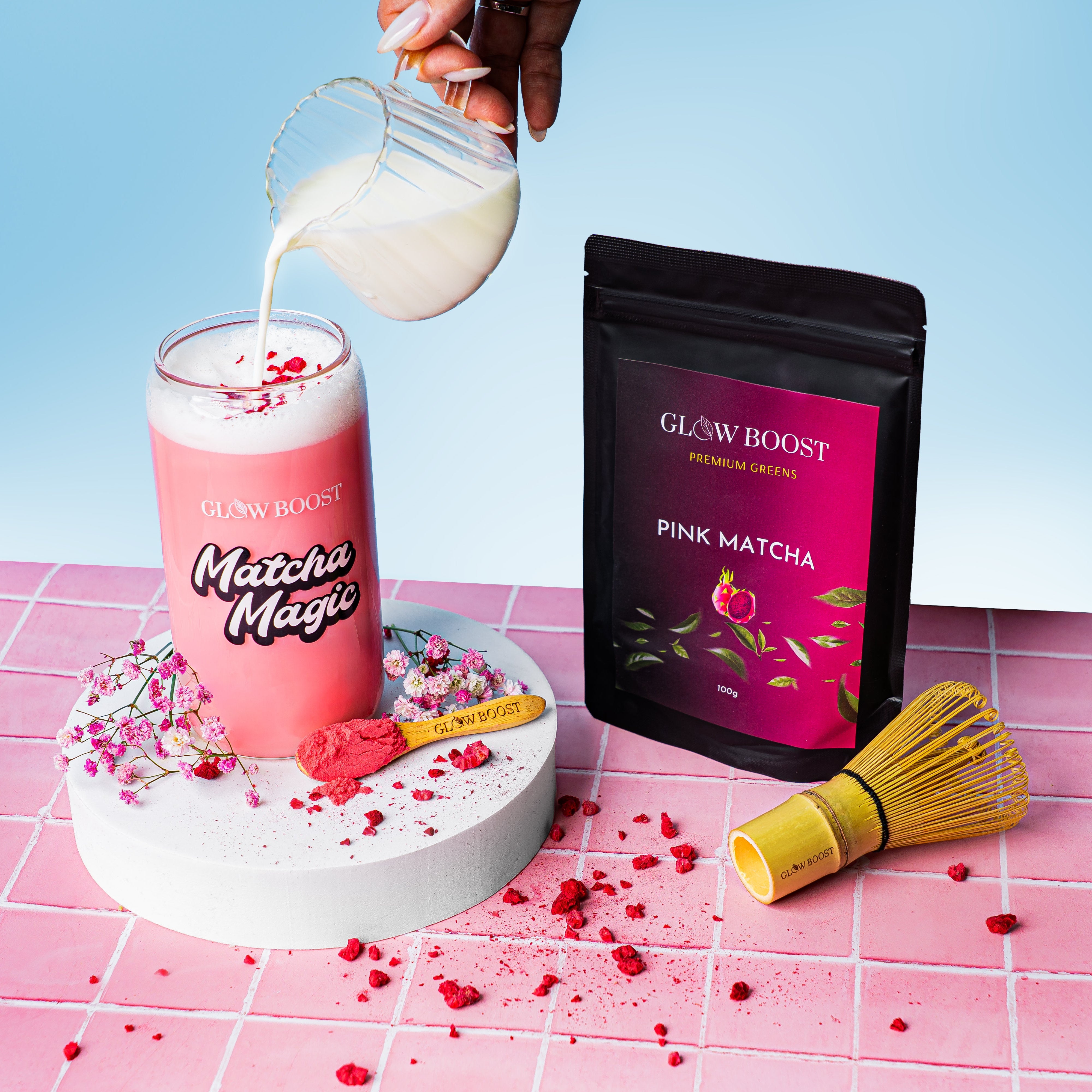 Pink matcha latte being poured into a glass with a package of GLOW BOOST Pink Matcha on a pink and blue background.